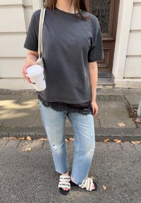Tricou din bumbac gri închis peste un top negru cu dantelă, blugi deschiși la culoare, uzati, și sandale negre cu curea crem. Ține o cană de cafea.