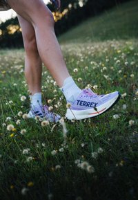 Pantaloni sportivi viola con una tomaia in tessuto testurizzato, audace marchio "TERREX" e suola ammortizzata, abbinati a calzini bianchi alla caviglia, in un campo erboso.