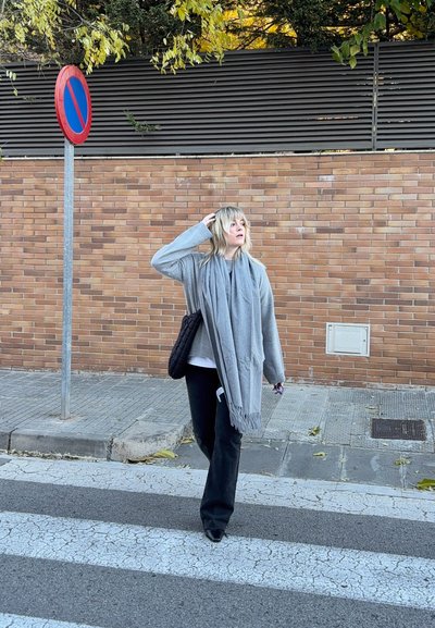 Mujer con bufanda y suéter grises, pantalones negros, llevando una bolsa negra, caminando por un paso de peatones cerca de una señal de no estacionar junto a una pared de ladrillos.