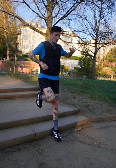 Chaleco negro, camiseta azul, pantalones cortos negros y zapatillas de correr negras con rayas blancas. Tatuajes visibles en las piernas. Corriendo por las escaleras al aire libre.