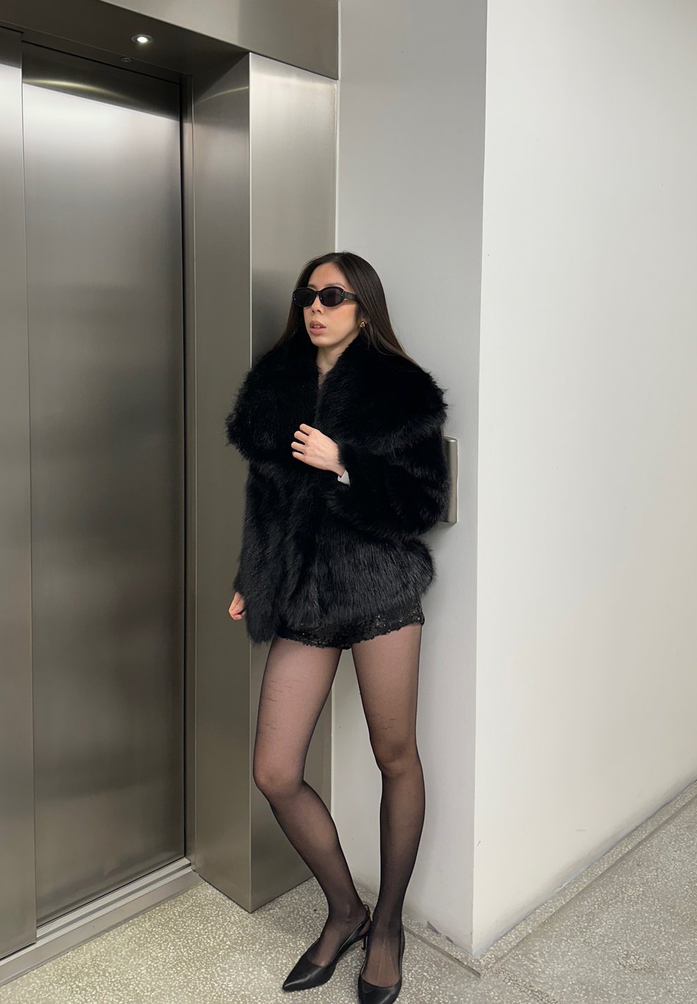 Manteau en fausse fourrure noire avec un large col, associé à un short noir pailleté, des collants noirs transparents et des talons noirs pointus. Décor moderne et épuré.
