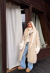 Personne portant un manteau en fourrure beige, une capuche en tricot blanc, des moufles et un jean bleu, se tenant près de l'entrée d'une cabane en bois avec des rideaux blancs.