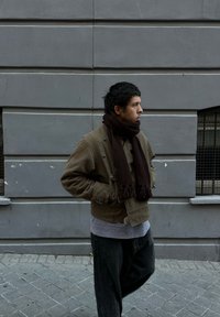 Üle suur pruuni jope nööpidega, allpool hall kampsun, tumedad laiad püksid ja tumepruun sall, halli seina taustal.