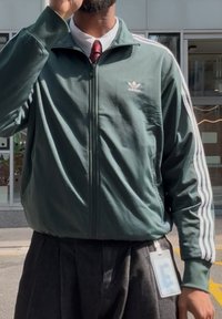 Temnozelena Adidas jakna na zadrgo z belimi črtami na rokavih, nosi se čez sivo hlače. Na levem prsnem predelu ima majhen logotip.
