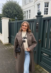 Jeune femme en manteau marron et jeans clairs, souriante, debout sur un trottoir à côté d'un portail vert foncé et d'un bâtiment blanc.