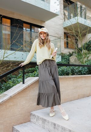 Suéter de punto amarillo claro, falda larga de capas verde oliva y bailarinas amarillas. Sosteniendo un libro, de pie en escalones exteriores. Follaje verde de fondo.