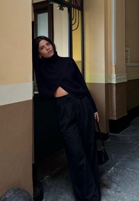 Suéter negro de punto corto con cuello alto, combinado con pantalones negros de talle alto. El fondo presenta una entrada con acentos de vidrio amarillo.