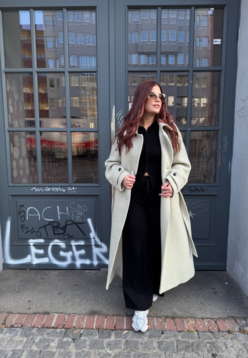 Gai pelēks vilnas mētelis ar lielām pogām, uzvilkts virs melna tērpa. Cilvēkam ir gaismas sporta apavi un apaļas saulesbrilles.