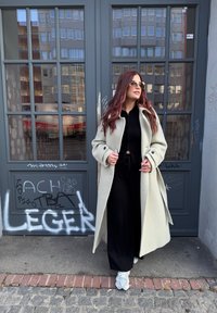 Gai pelēks vilnas mētelis ar lielām pogām, uzvilkts virs melna tērpa. Cilvēkam ir gaismas sporta apavi un apaļas saulesbrilles.