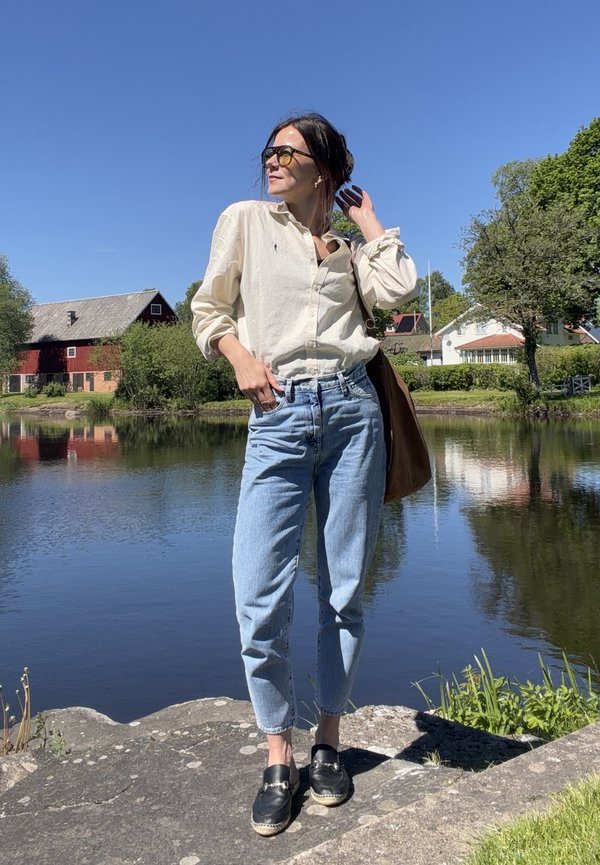 Vrouw met zonnebril, beige blouse, blauwe spijkerbroek en zwarte schoenen staand op rotsen bij een kalm meer met huizen en bomen op de achtergrond.