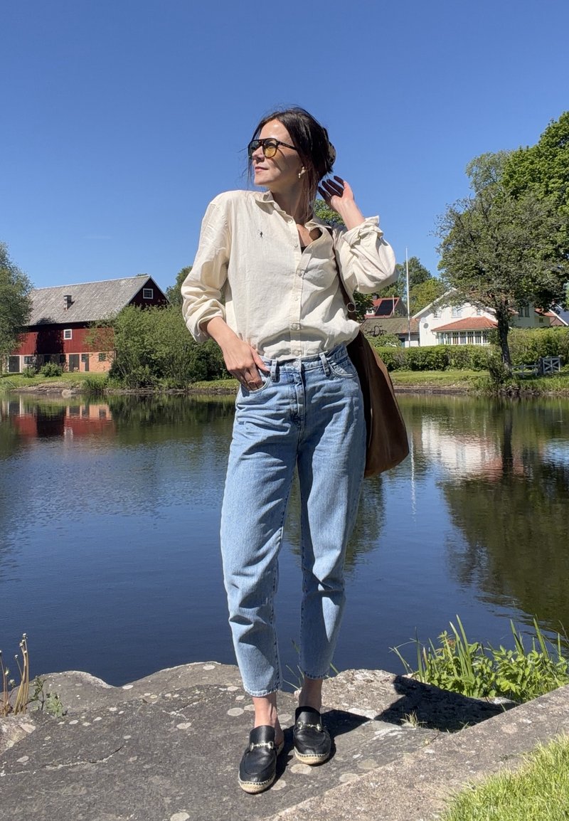 Femme portant des lunettes de soleil, une chemise beige, un jean bleu et des chaussures noires debout sur des rochers au bord d'un lac calme avec des maisons et des arbres en arrière-plan.