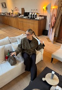 Femme aux cheveux tressés et portant des lunettes, assise sur un canapé blanc, tenant un sac à main dans une pièce moderne avec des machines à café et un grand portrait.