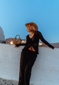 Top lungo a maniche nere in pizzo con coulisse, abbinato a pantaloni neri larghi. Un cappello di paglia e una borsa rotonda intrecciata con manico completano l'outfit.
