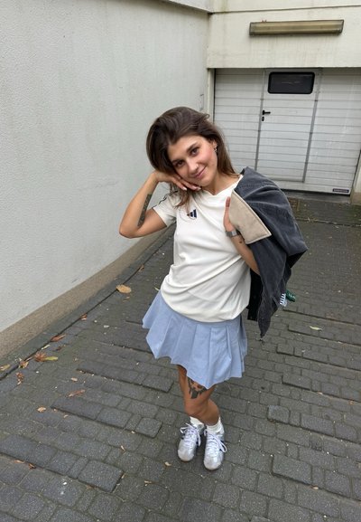 Camiseta ligera de crema con logo, falda plisada azul clara, chaqueta de mezclilla gris, zapatillas plateadas, tatuaje estampado en la pierna izquierda.