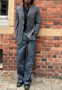 Blazer gris sobre una camisa negra, combinado con pantalones vaqueros azules sueltos. Zapatos negros completan el atuendo. El fondo presenta una pared de ladrillo.