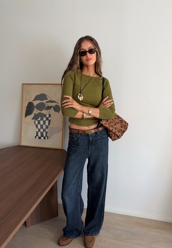 Mujer con cabello largo que lleva gafas de sol, camiseta de manga larga verde oliva, vaqueros de pierna ancha, bolso con estampado de leopardo, de pie junto a una mesa de madera con una obra de arte de planta enmarcada.