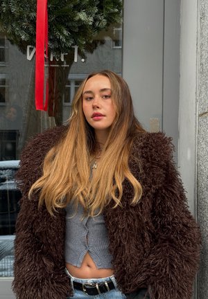 Joven con cabello rubio largo que lleva una chaqueta esponjosa marrón oscuro y un top gris recortado, de pie frente a una puerta de vidrio decorada.