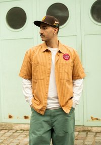 Korte mouwen, oranje gestreept shirt met twee voorzakken, gelaagd over een wit shirt met lange mouwen. Olijfgroene broek en een bruine pet.