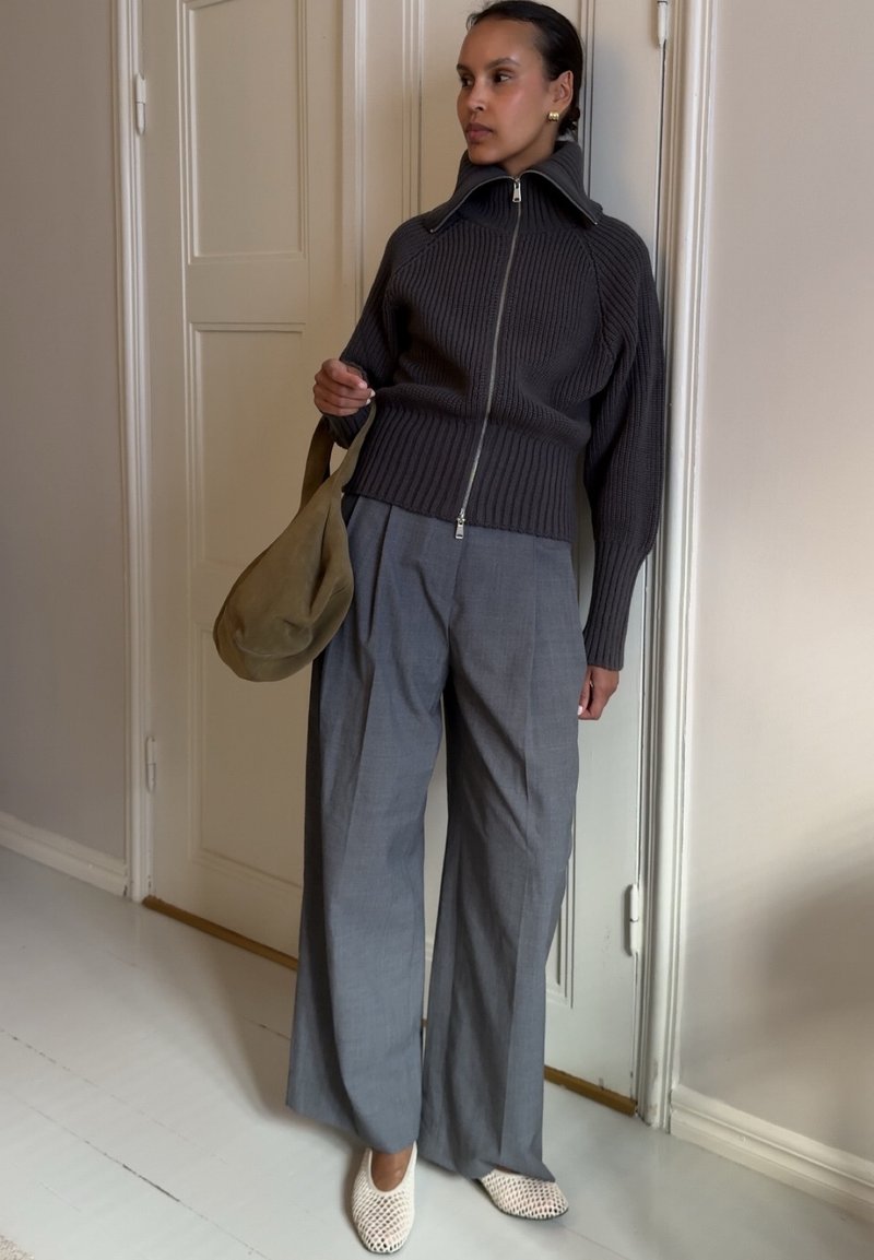 Femme appuyée contre une porte, portant un pull zippé foncé, un pantalon large gris, des chaussures blanches tissées et tenant un sac vert olive.