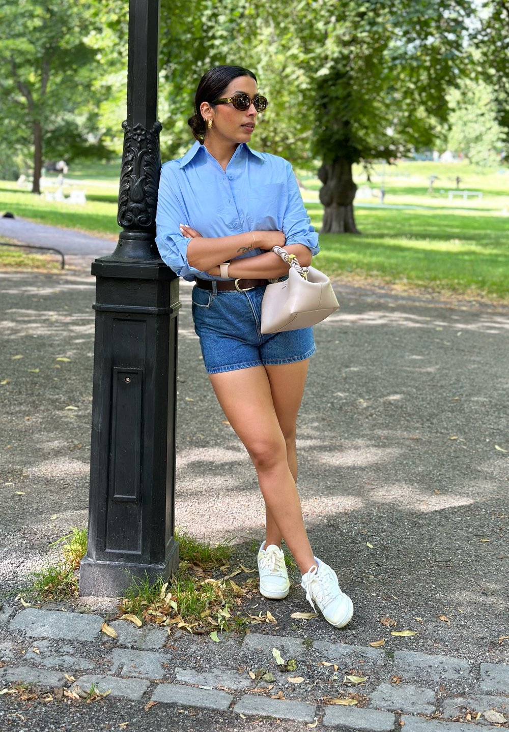 Camisa de botones azul claro, pantalones cortos de mezclilla y zapatillas blancas. Sosteniendo un bolso beige, apoyada contra una farola negra en un parque verde.