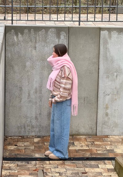 Mujer con suéter a rayas, bufanda rosa, jeans anchos azules y zapatos beige, de pie de lado sobre un camino de ladrillos frente a una pared de concreto.