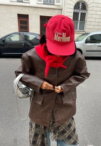 Hnědá kožená bunda s velkými kapsami, červený šátek a červená kordurová čepice s bílým textem. Stříbrná kabelka s řetízkovým popruhem.