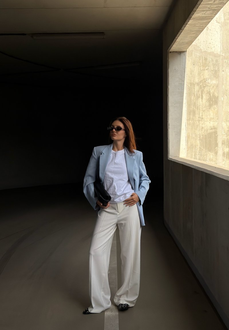 Mujer con blazer azul claro, camiseta blanca y pantalones anchos blancos, de pie con confianza en un aparcamiento oscuro cerca de una gran ventana de hormigón, con gafas de sol.