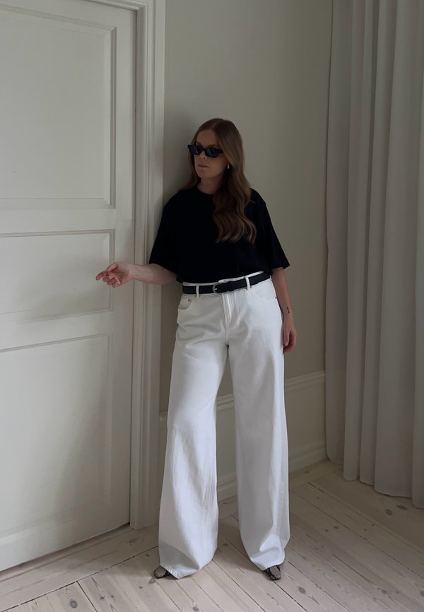 Femme aux longs cheveux bruns portant des lunettes de soleil noires, une chemise noire, un pantalon blanc à jambes larges et des chaussures à motifs, debout près d'une porte à l'intérieur.