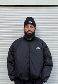 Sort bomberjakke med lynlås foran og sidelommer, med et logo på brystet. Båret sammen med en matchende sort strikket beanie. Ensfarvet baggrund.