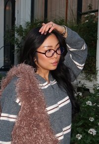 Grijze trui met witte en kastanjebruine strepen, gecombineerd met een pluizige, lichtroze sjaal. Ronde bril en lang, donker haar maken de look compleet.