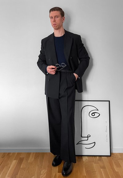 Hombre con traje negro y camisa azul marino de pie sobre suelo de madera sosteniendo unas gafas junto a un cuadro con arte minimalista de líneas faciales sobre una pared gris.