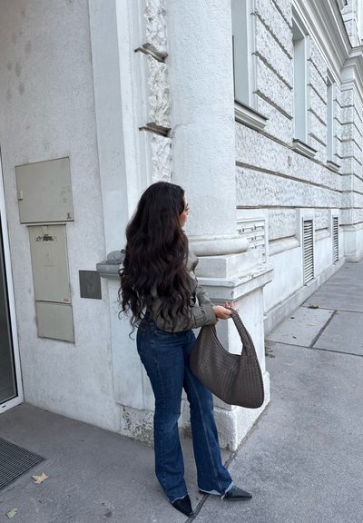 Una mujer con una chaqueta acolchada verde y jeans acampanados azules está de pie sosteniendo un bolso tejido marrón, con el cabello largo y ondulado, frente a una pared texturizada.