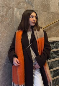 Jeune femme aux cheveux bruns mi-longs portant un manteau noir et une écharpe orange-beige, debout près d'un mur en marbre à côté d'une rampe d'escalier.