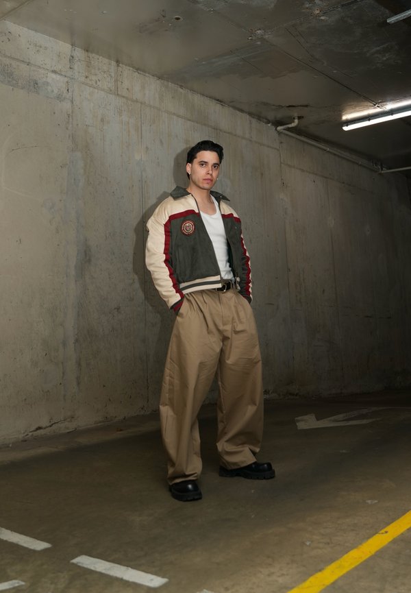 Joven está de pie en un aparcamiento de hormigón tenue, vistiendo pantalones holgados kaki, camiseta blanca sin mangas y chaqueta con bloques de color, con las manos en los bolsillos.