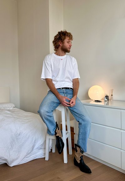 Camiseta blanca de manga corta con un pequeño logo rojo, jeans de color azul claro y botas hasta el tobillo de color negro-marrón. Sentado en un taburete blanco en una habitación sencilla.