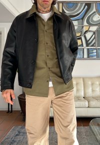 Veste en cuir noire avec col, portée sur une chemise boutonnée vert olive, associée à un pantalon beige. En arrière-plan, un canapé clair et une œuvre d'art.