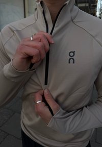 Személy bézs cipzáros dzsekit visel, amelyen logó látható, részben lecipzározza azt, bemutatva ezüst gyűrűkkel díszített kezét.