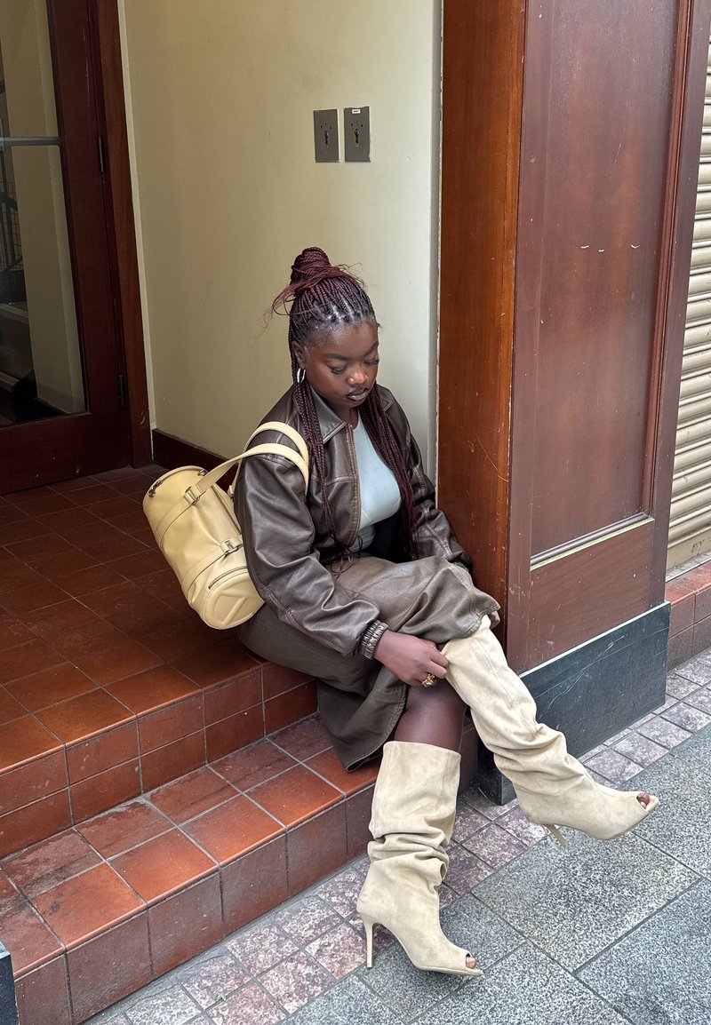 Giovane donna che sistema stivali beige con tacco alto mentre è seduta su gradini piastrellati all'aperto, indossando una giacca di pelle marrone e portando uno zaino beige.