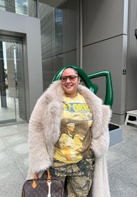 Γυναίκα που χαμογελά φορώντας μεγάλο μπεζ παλτό από γούνα, κίτρινη γραφική μπλούζα, παντελόνι καμουφλάζ και γυαλιά ηλίου, κρατώντας ένα καφέ δερμάτινο τσάντα σχεδιαστή σε εξωτερικό χώρο.