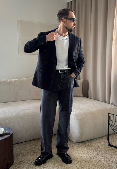 Hombre con blazer oscuro, camiseta blanca, vaqueros oscuros, zapatos negros y gafas de sol, de pie en una sala de estar con sofá beige y cortinas.