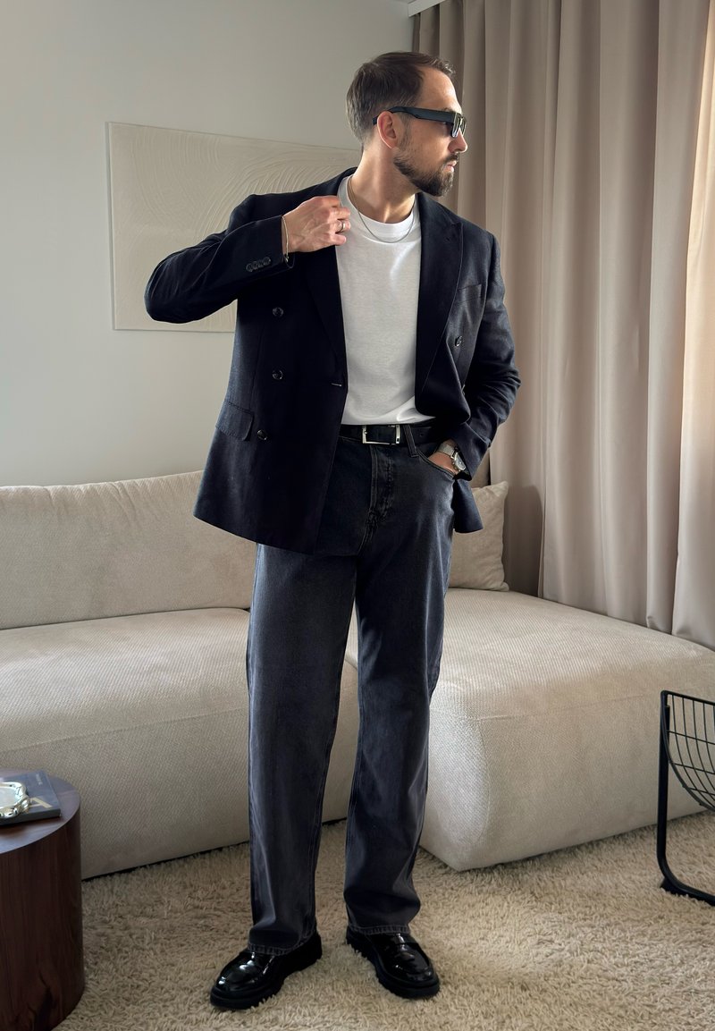Homme portant un blazer foncé, un t-shirt blanc, un jean foncé, des chaussures noires et des lunettes de soleil, debout dans un salon avec un canapé beige et des rideaux.