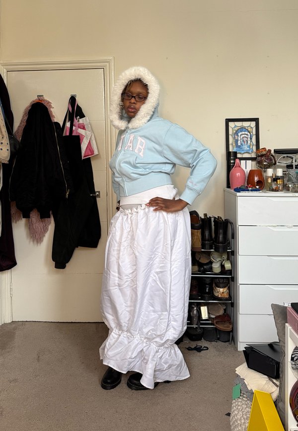 Személy világoskék kapucnis kabátban, melynek szegélyét szőrme díszíti, és hosszú fehér fodros szoknyában áll egy szobában, amely tele van kabátokkal, cipőkkel és egy komód található benne.
