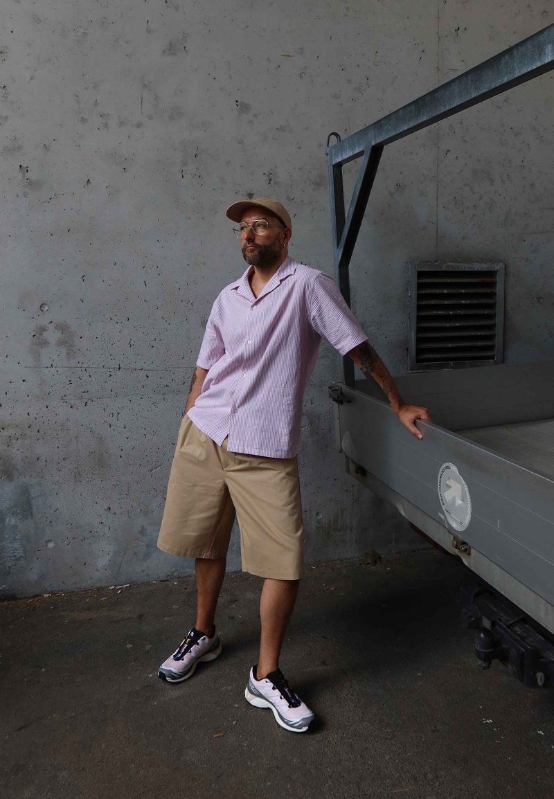Homme portant une casquette beige, des lunettes, une chemise rose rayée et un short kaki, appuyé contre un véhicule gris devant un mur en béton.