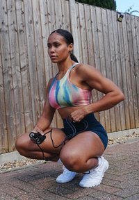 Top cropped desportivo em padrão tie-dye pastel; calções pretos de cintura alta; sapatilhas brancas. A segurar uma corda de saltos enquanto está agachada.