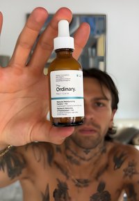Man met tatoeages die een bruine pipetfles met het etiket "The Ordinary Skin Care" dicht bij de camera houdt in een kamer met een ingelijste foto.