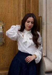 Vrouw met lang donker haar in een witte blouse en een donkere rok staat bij een houten deur, haar nek aanraken en naar beneden kijken, leunend tegen een stenen muur.
