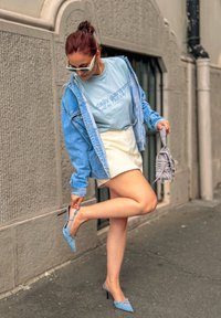 Giacca di jeans, t-shirt grafica azzurro chiaro, mini gonna bianca e décolleté azzurro chiaro. Scarpe grigie tenute in mano, sfondo urbano.
