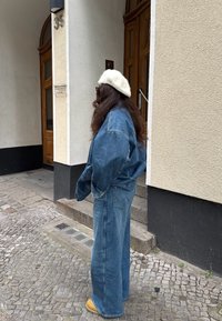 Hele sinine teksajakk ja laiad teksapüksid, millele on lisatud valge tehiskarvberet. Väljasett, kus on kiviparkett ja puidust uks.