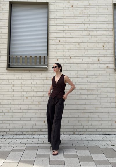 Mujer con top sin mangas oscuro, pantalones anchos negros, gafas de sol y sandalias, de pie junto a una pared de ladrillo clara con ventana cerrada.