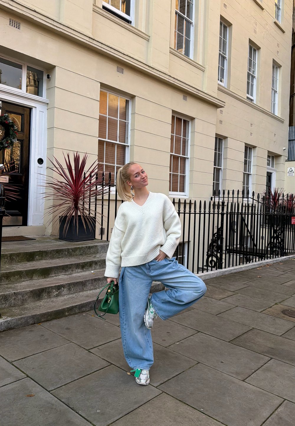 Pull blanc oversize, jeans bleu à jambes larges, sac à main vert et baskets argentées. En extérieur avec un bâtiment et des plantes décoratives.